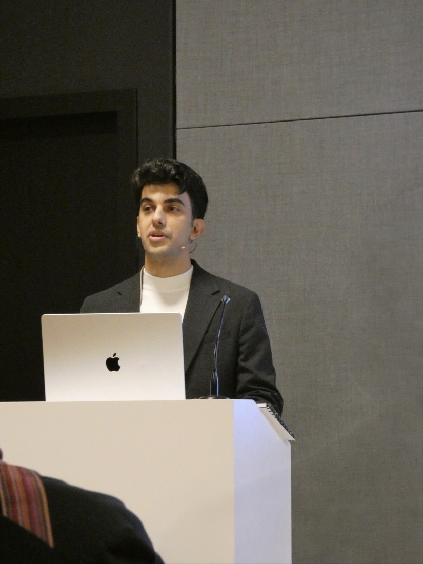 Ashkan Pirmani speaking at a white lectern with a laptop open in front of him
                      and a headset microphone on.