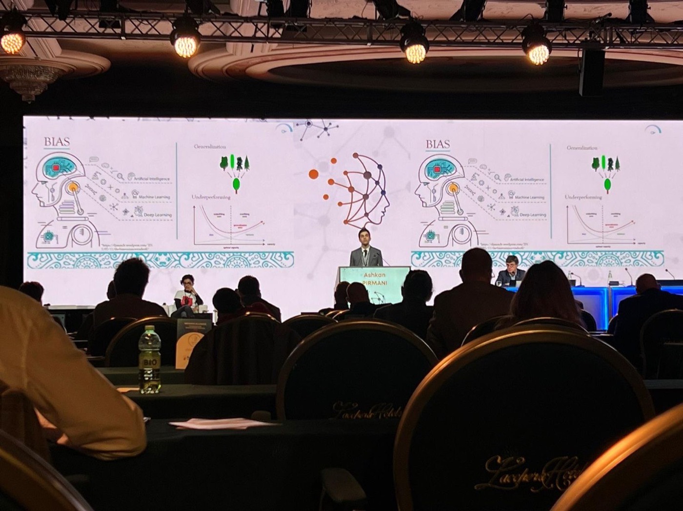 A large conference stage. Ashkan Pirmani stands at a lectern in front of a wide
screen showing slides about bias and generalization. Several hundred people are
seated in front of him.